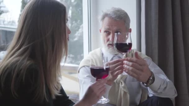 Handsome senior man with wineglass in hand talking to young brunette Caucasian woman indoors. Carefree elegant retiree spending evening with younger partner. Age difference, relationship, love. — Stock Video
