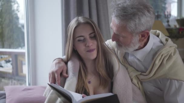 Homem caucasiano sênior apaixonado conversando com a jovem mulher branca bonita ler livro. Feliz casal amoroso com diferença de idade desfrutando de tempo juntos dentro de casa. Estilo de vida, amor, romance, tranquilidade . — Vídeo de Stock