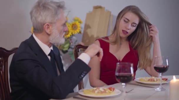 Retrato de jovem mulher sedutora em vestido vermelho sentado com o homem sênior na mesa e sorrindo. Mulher encantadora de virtude fácil flertando com empresário de sucesso durante o jantar romântico . — Vídeo de Stock