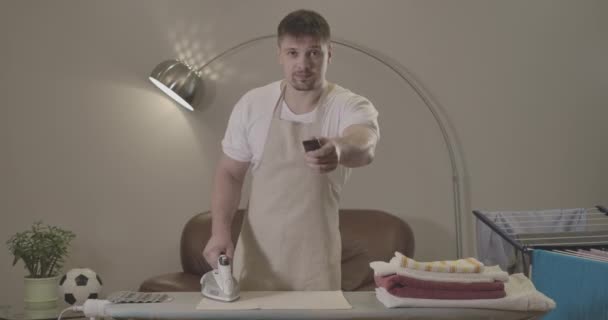 Retrato de joven guapo morena hombre cambiando canales como de pie delante de la tabla de planchar. Adulto caucásico ama de llaves haciendo tareas domésticas y viendo la televisión. Sede del cine 4k ProRes . — Vídeos de Stock