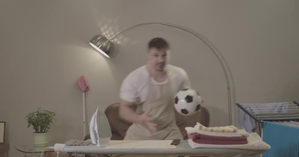 Actieve jonge blanke man met voetbal juichen als kijken naar wedstrijd op TV binnen. Opgewonden knappe man die een overwinningsgebaar maakt voor de strijkplank met de was. Bioscoop 4k Prores Hq. — Stockvideo