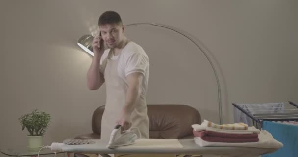 Portrait of young Caucasian man talking on the phone and ironing laundry. Brunette handsome guy dealing with housekeeping and chatting. Household, lifestyle, joy. Cinema 4k ProRes HQ. — Stock Video