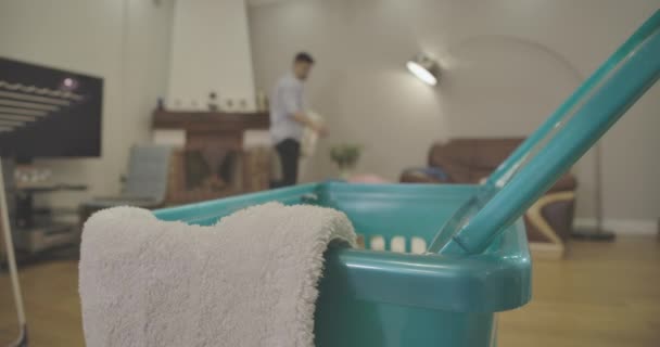 Close-up of household equipment standing on foreground as blurred young Caucasian man gathering dirty laundry at the background and putting in into basket. Housekeeping, lifestyle. Cinema 4k ProRes HQ — 图库视频影像