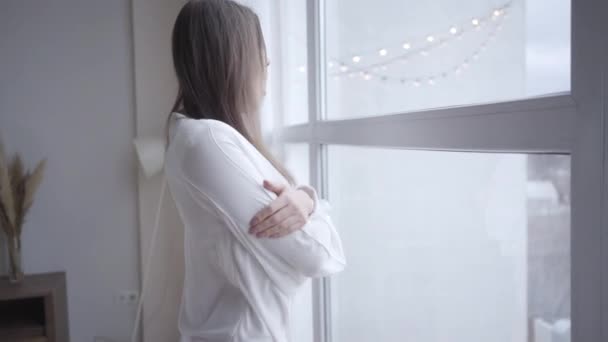 Närbild av sömnig ung vit kvinna gäspande och tittar ut genom fönstret på morgonen. Sidovy över söt brunett flicka vaknar hemma. Livsstil, fritid, glädje. — Stockvideo