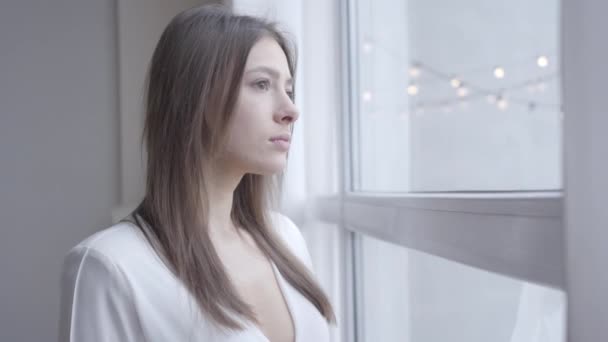 Närbild av omtänksam brunett vit kvinna med bruna ögon tittar ut genom fönstret hemma. Söt tjej som vaknar på morgonen på helgerna. Livsstil, glädje, fritid. — Stockvideo