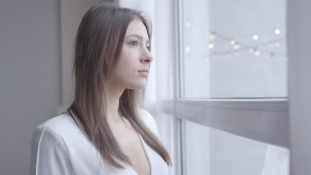 Primer plano de la morena reflexiva mujer caucásica con ojos marrones mirando por la ventana en casa. Linda chica despertando por la mañana los fines de semana. Estilo de vida, alegría, ocio . — Vídeo de stock