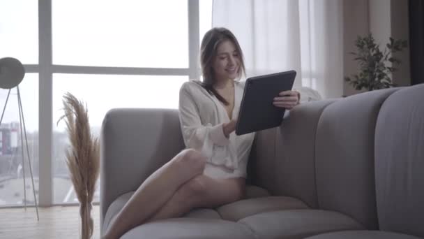 Bájos mosolygós fehér lány ül a kanapén, és a tabletta. Portré egy vidám fiatal nőről, aki a közösségi médiát használja reggel. Joy, életmód, vezeték nélküli technológia. — Stock videók