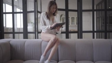 Tablet kullanan güzel beyaz bir kadının portresi kanepede oturuyor. Sabahları sosyal medyayı kontrol eden esmer bir kız. Yaşam tarzı, internet, kablosuz teknoloji.
