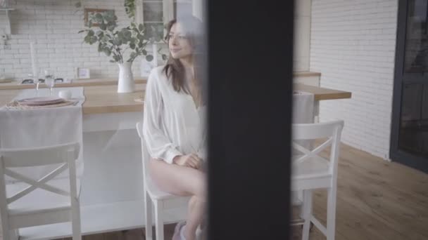 Joven encantadora morena caucásica sentada en la cocina y cruzando las piernas. Sonriendo hermosa chica disfrutando de la mañana tranquila en el interior. Disparando a través de cristal y marco de la puerta. Ocio, estilo de vida, alegría . — Vídeos de Stock