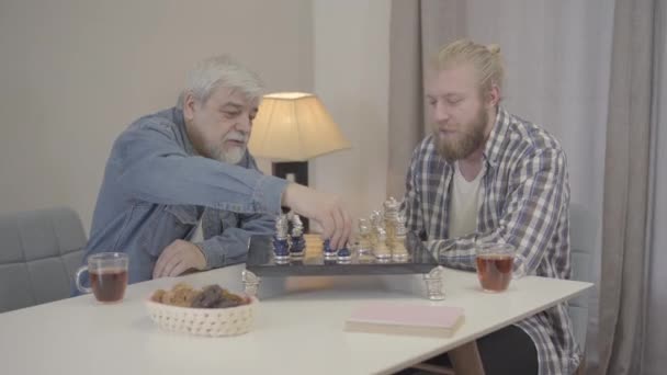 İçeride satranç oynayan genç ve yaşlı adamların portresi. Kafkas yetişkin oğlu olgun babasıyla mutlu bir akşam geçiriyor. Boş zaman, yaşam tarzı, birlik, aile. — Stok video