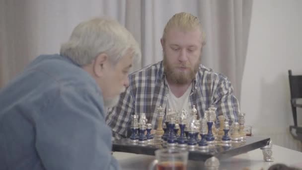 A fiatal, fehér szakállas férfi portréja az idősebb apával való játékra koncentrált. Szürke hajú nyugdíjas férfi sakkozik a felnőtt fiával hétvégenként otthon. Szabadidő, hobbi, életmód. — Stock videók
