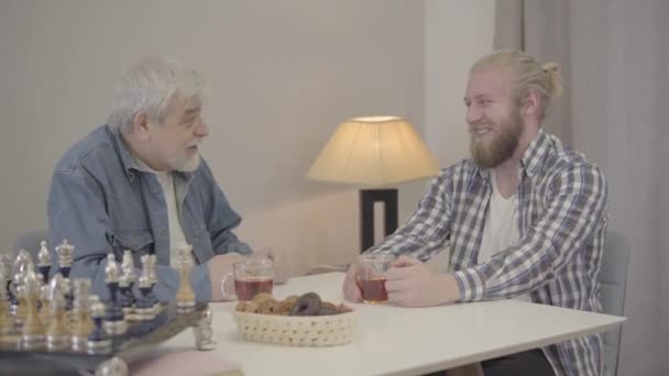 Двоє веселих кавказьких чоловіків сміються і п'ють чай у приміщенні. Щасливий молодий син і старший батько відпочивають вдома на вихідних. Радість, спосіб життя, дозвілля, щастя. — стокове відео