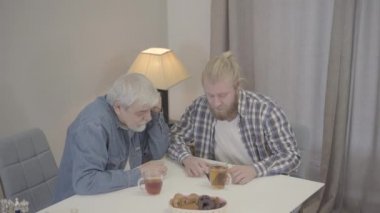 Olumlu Kafkasyalı yetişkin oğul ve olgun baba birlikte müzik dinliyorlar. Gülümseyen adamlar kulaklık takar ve akıllı telefondan şarkılar açarlar. Mutluluk, birlik, yaşam tarzı.