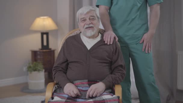 Portret van een oudere blanke man die voor de camera praat als een onherkenbare mannelijke verpleegster die hand in hand op zijn schouder houdt. Positieve gepensioneerde brengt een rustige avond door in het verpleeghuis. Zorg, veroudering, levensstijl, hulp. — Stockvideo