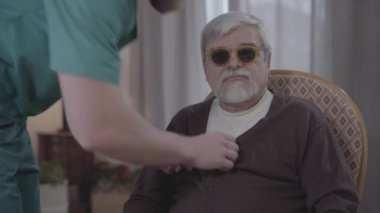 Evde oturan kör beyaz adamın portresi ceketinin düğmesini bağlayan erkek hemşire gibi ve gidiyor. Yetişkin bir yardımcı huzurevinde olgun bir emekliye bakıyor. Yaşam tarzı, yardım, destek.