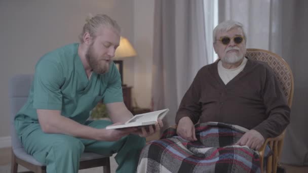 Mężczyzna biały ochotnik w mundurze pielęgniarki czyta książkę dla niewidomych emerytów w domu. Współczujący mężczyzna spędzający wieczór z dojrzałym emerytem w domu opieki. Styl życia, opieka, wsparcie, pomoc. — Wideo stockowe
