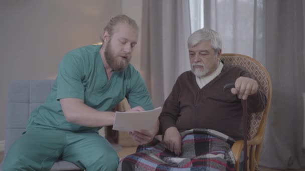 Retrato de enfermeira masculina grave em documentos de leitura uniformes para o velho homem caucasiano sentado em poltrona com bengala. Jovem assistente médico ajudando aposentado para lidar com papéis. Apoio, cuidado, envelhecimento . — Vídeo de Stock