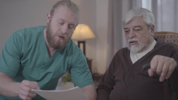 Közelkép a fehér férfi ápolóról, aki az érett nyugdíjasoknak magyarázza a dokumentumokat. Az öregek otthonában asszisztál. Segítség, támogatás, öregedés, életmód. — Stock videók