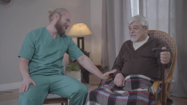 Hombre caucásico adulto positivo en uniforme de enfermera charlando con un jubilado varón maduro en un asilo de ancianos. Joven barbudo y pensionista mayor sonriendo y riendo. Cuidado, estilo de vida, apoyo, envejecimiento . — Vídeo de stock