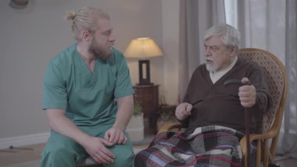 Portrait of Caucasian man listening to stories of upset retiree in nursing home and holding his hand. Male nurse supporting old man. Care, help, volunteering, lifestyle. — Stock Video
