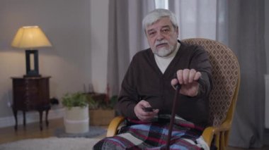 Yürüyen baston kanalı uzaktan kumandayla değiştiren yaşlı beyaz adamın portresi. Emekli yaşlı erkek emeklilik maaşıyla evde dinleniyor. Yaşam tarzı, rahatlık, rahatlama, yaşlanma..