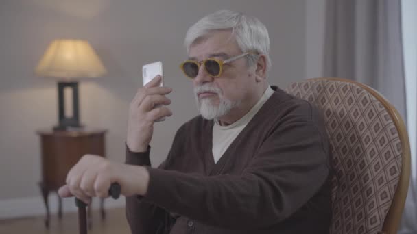 Portret van een blinde oude blanke man die binnen aan het bellen was. Grijsharige man gepensioneerd in fauteuil met smartphone en chatten. Moderne technologieën voor gehandicapten, levensstijl. — Stockvideo