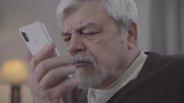 Közelkép egy szomorú szemű öregemberről, aki okostelefon képernyőt lop. Az öreg fehér nyugdíjas telefonál idebent. Modern technológiák, életmód, öregedés, rövidlátás. — Stock videók