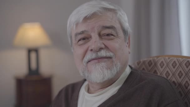 Zbliżenie siwego białego mężczyzny z brązowymi oczami uśmiechniętymi do kamery. Portret szczęśliwego emeryta pozującego w domu. Radość, styl życia, emerytura, emerytura. — Wideo stockowe