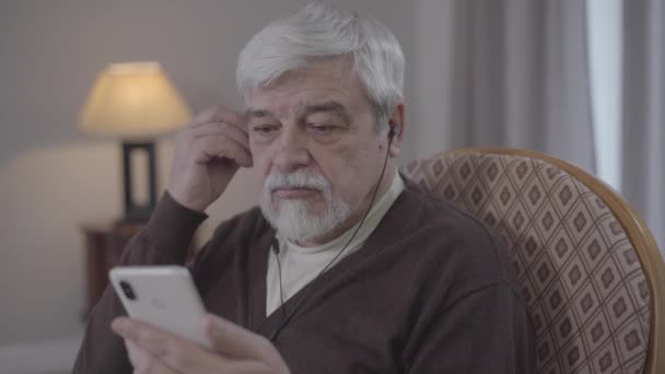 Detailní záběr zralého bělocha, jak si nasazuje sluchátka a zapíná hudbu na smartphonu. Pozitivní důchodce, který doma poslouchá playlist. Hobby, štěstí, životní styl, odchod do důchodu. — Stock video