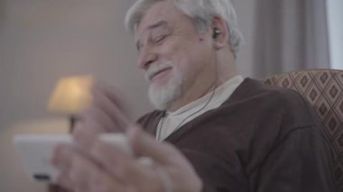 Kulaklık takmış, akıllı telefondan film izleyen kıdemli beyaz adamın yakın çekimi. Akşamını evde geçiren neşeli, olgun bir emeklinin portresi. Kablosuz teknoloji, yaşam tarzı, yaşlanma.