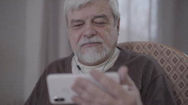 Kulaklık takıp akıllı telefondan film izleyen kahverengi gözlü son sınıf öğrencisinin yüzü. Duygusal tepki veren ve gülümseyen mutlu emeklilerin portresi. Yaşam tarzı, yaşlanma, rahatlama, rahatlama..