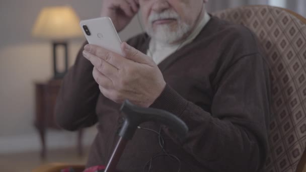 Неузнаваемый старик включает плейлист на смартфоне и слушает музыку в наушниках. Положительный зрелый пенсионер наслаждается свободным временем на пенсию. стиль жизни, счастье, хобби, досуг . — стоковое видео