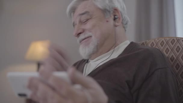 Gros plan d'un homme caucasien âgé riant dans des écouteurs regardant des séries de films sur smartphone. Portrait de retraité mature joyeux passant la soirée à l'intérieur. Technologie sans fil, mode de vie, vieillissement . — Video