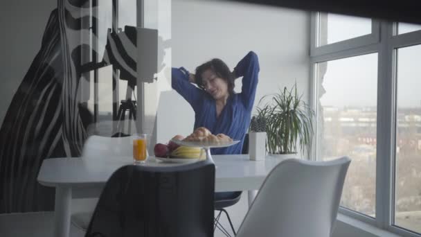 Студийный кадр счастливой взрослой белой женщины, завтракающей утром в помещении. Улыбающаяся брюнетка растягивается, как сидя за столом с соком и фруктами. Видеооборудование в отражении . — стоковое видео