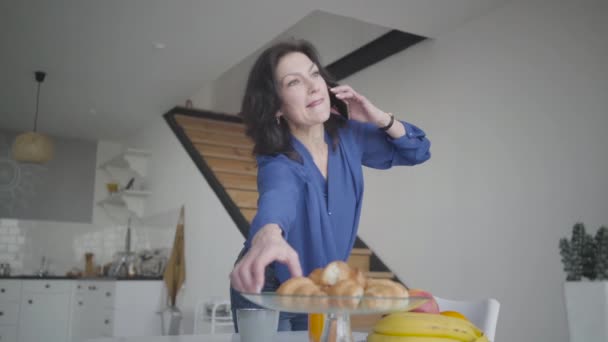 Donna d'affari caucasica di successo che parla al telefono durante la colazione leggera a casa. Ritratto di donna bruna sicura che prende croissant e bicchiere di succo d'arancia durante la conversazione. Stile di vita . — Video Stock