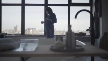 Kahve içen ve büyük pencerenin önünde gerinen muhteşem beyaz kadın portresi. Mutlu başarılı iş kadını evde eğleniyor. Zenginlik, lüks, yaşam tarzı.