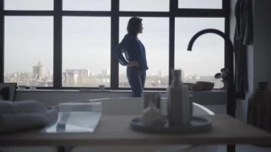 Esmer, beyaz bir kadının pencereden dışarı bakıp pencere eşiğine oturduğu uzun bir çekim. Düşünceli muhteşem bayan sabahını evde geçiriyor. Yaşam tarzı, konfor, rahatlık.