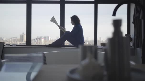 Mujer de negocios caucásica seria sentada en el alféizar de la ventana y leyendo el periódico. Retrato de vista lateral de una mujer morena adulta tomando café y examinando noticias por la mañana. Éxito, estilo de vida, negocios . — Vídeo de stock