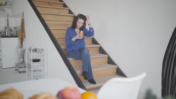 Plan large de l'élégante femme brune caucasienne buvant du jus d'orange le matin à la maison. Portrait de belle dame confiante assise sur les escaliers à l'intérieur. Mode de vie, luxe, richesse, richesse . — Video