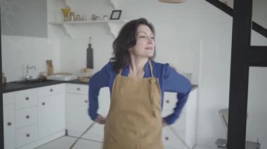 Önlük giyip kameraya gülümseyen çekici esmer beyaz kadın. Mutfakta poz veren güzel bir ev kadınının portresi. Yaşam tarzı, mutluluk, ev hayatı..