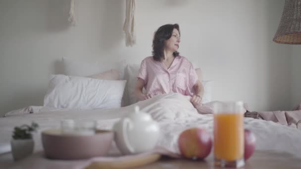 Retrato de una mujer caucásica morena en pijama sentada en la cama por la mañana. Señora positiva disfrutando de fin de semana en casa. Jugo orgánico fresco, té y frutas en primer plano. Estilo de vida, ocio . — Vídeo de stock