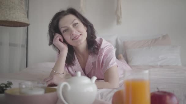Positieve brunette Kaukasische vrouw in roze pyjama kijkend naar camera en glimlachend. Portret van een vrolijke vrouw die 's ochtends in de slaapkamer doorbrengt. Vreugde, vrije tijd, levensstijl, geluk. — Stockvideo