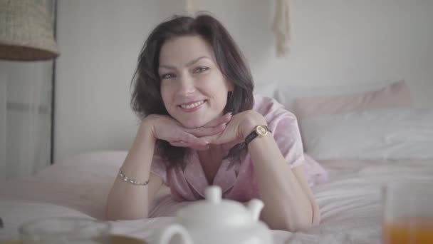 Портрет позитивної кавказької жінки-брюнетки, що позувала вранці в спальні. Гарна молода леді лежить на ліжку і посміхається. Відпочинок, відпочинок, вихідні, спосіб життя. — стокове відео