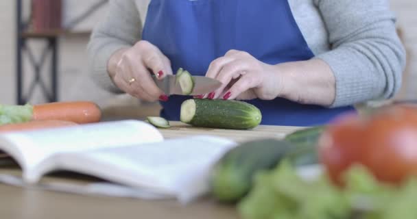 Close-up de feminino caucasiano mão sênior cortando pepino orgânico. Mulher de meia-idade irreconhecível preparando salada saborosa fresca em casa. Culinária, culinária, estilo de vida. Cinema 4k ProRes HQ . — Vídeo de Stock