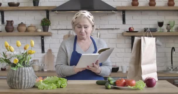 Femme âgée caucasienne positive à la recherche d'une nouvelle recette de salade debout dans la cuisine. Portrait de belle femme au foyer d'âge moyen lisant un livre de recettes, légumes couchés sur la table. Siège social Cinema 4k ProRes . — Video