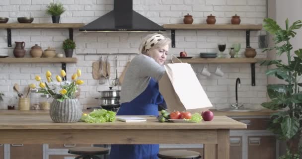 Portrét pozitivní kavkazské starší ženy, která vytahuje mrkev a okurky z nákupní tašky. Krásná středního věku důchodce vaření v moderní kuchyni uvnitř. Cinema 4k ProRes HQ. — Stock video