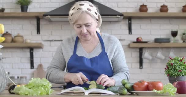 Sarışın, kıdemli, beyaz bir kadın salatalık kesip bir dilimi tadıyor. Mutlu ev hanımı portresi salata pişiriyor ve kameraya gülümsüyor. Yaşam tarzı, sağlıklı beslenme, vejetaryenlik. Sinema 4k ProRes Merkezi. — Stok video