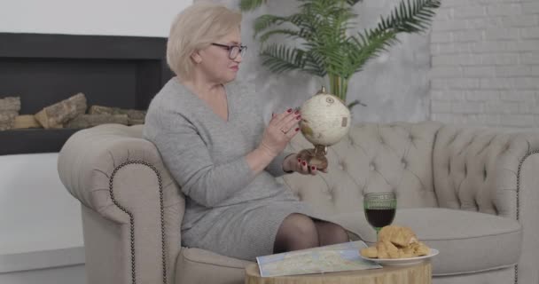 Side view of elegant Caucasian senior woman comparing earth globe and map. Middle-aged blond lady in eyeglasses deciding on vacations abroad. Tourism, travelling, lifestyle. Cinema 4k ProRes HQ. — Stock Video