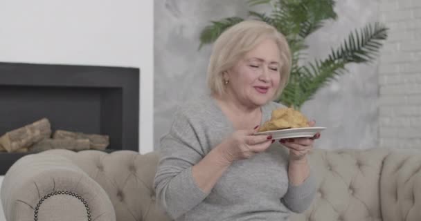 Retrato de mujer caucásica mayor feliz con ojos marrones oliendo y mordiendo delicioso croissant. El positivo rubio se retira desayunando ligero en casa. Riqueza, lujo, estilo de vida. Sede del cine 4k ProRes . — Vídeos de Stock