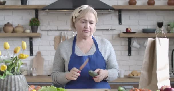 Mutfakta elinde salatalık ve bıçakla konuşan kendinden emin beyaz kadın portresi. Orta yaşlı emekli, evde yemek pişirmeyi açıklıyor. Yaşam tarzı, vloglama, aşçılık. Sinema 4k ProRes Merkezi. — Stok video